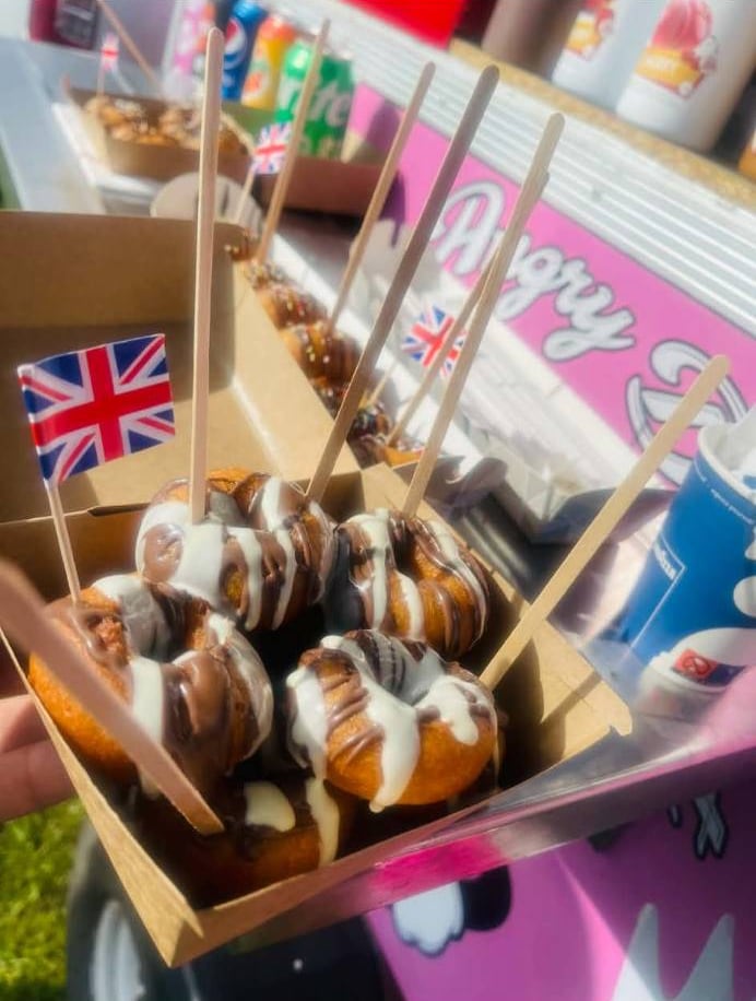 A box of fresh mini donuts drizzled with milk and white chocolate, served with wooden forks and a Union Jack flag