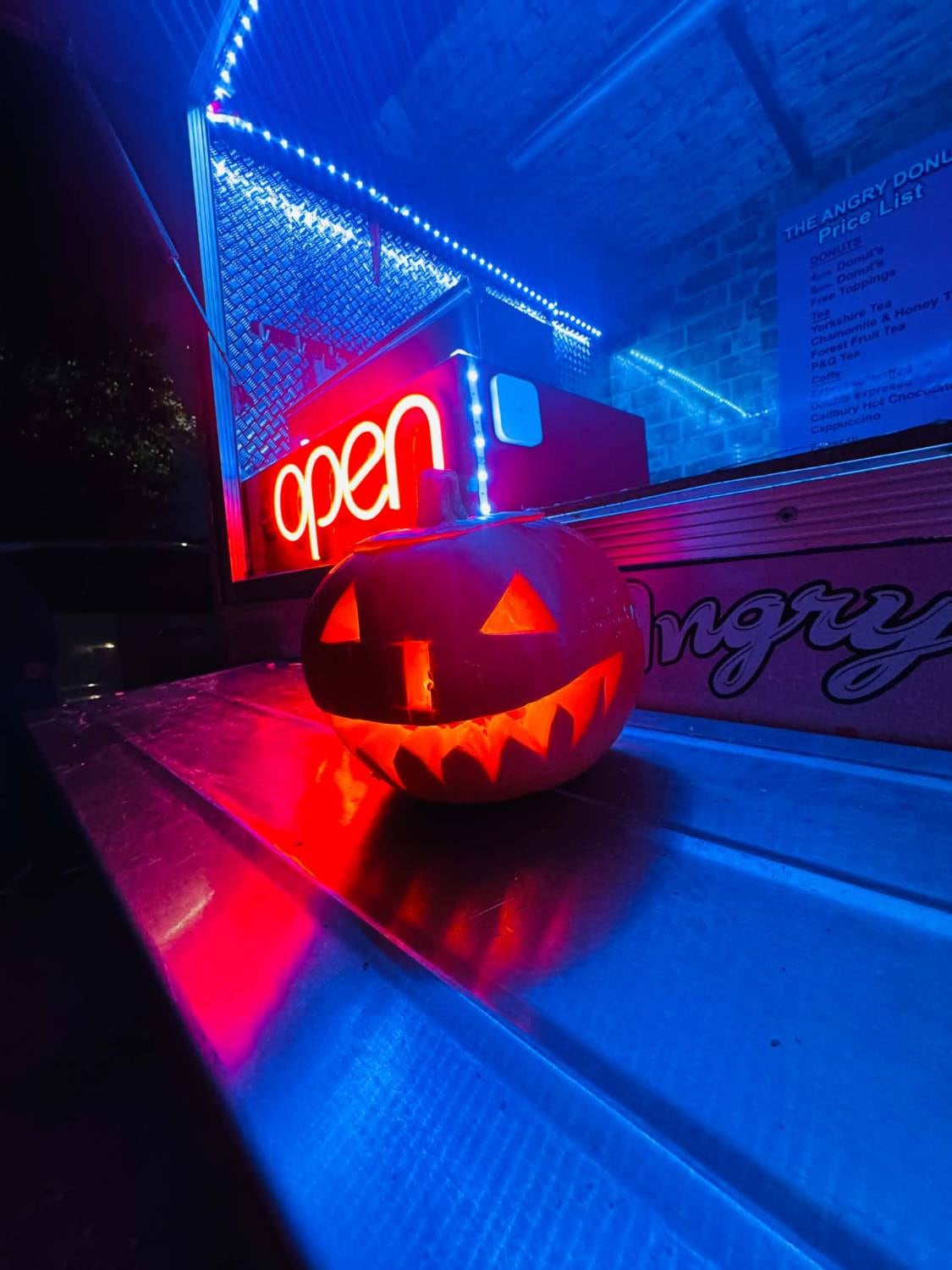 A carved pumpkin glowing on the Angry Donut counter beside the neon 'open' sign at night